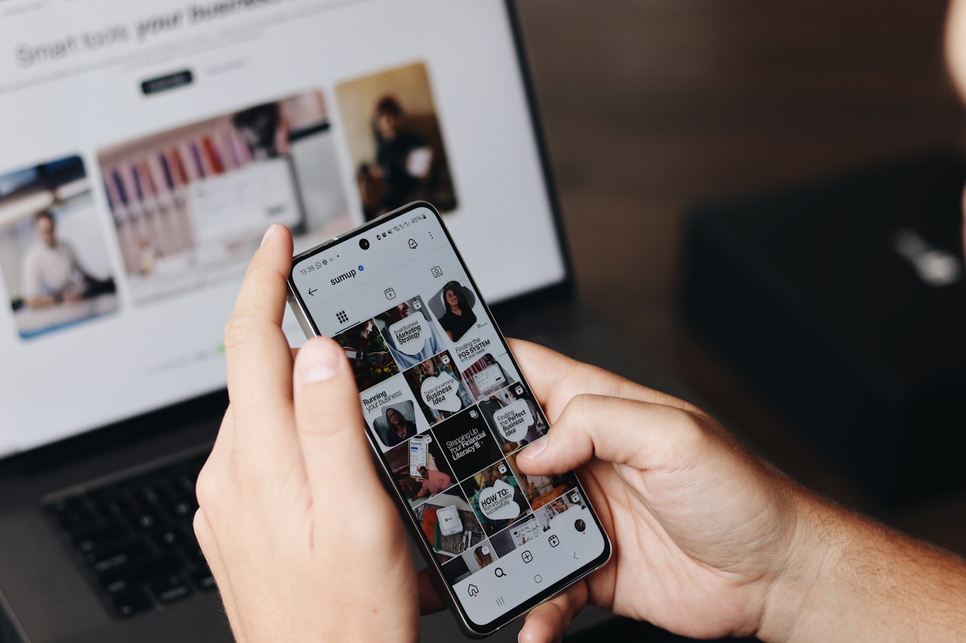 A creator holding a phone showing a grid of their content, with a laptop open behind it